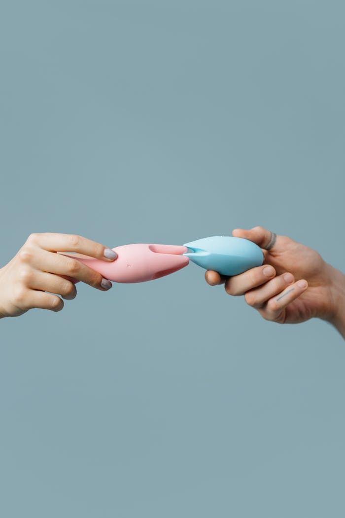 about-01 Two hands exchanging pink and blue silicone vibrators against a light background.